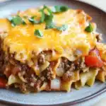 Beef casserole with ground beef and noodles - the image shows a plate of lasagna on a wooden table. the lasagna is made with long, thin noodles and topped with a layer of melted cheese. the cheese is golden brown and appears to be melted and bubbly. the top of the lasagna has a generous amount of ground beef, which is visible in the center. there are also chunks of red bell peppers and onions scattered throughout the dish. the plate is blue and has a speckled texture. the background is blurred, but it seems to be a kitchen countertop.
