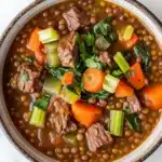 Beef lentil soup - the image is a close-up of a bowl of soup. the soup appears to be a beef stew with chunks of beef, carrots, celery, and other vegetables mixed in. the bowl is made of ceramic and is sitting on a white marble countertop. there are a few sprigs of parsley scattered around the bowl. the broth is a rich brown color and looks thick and hearty.