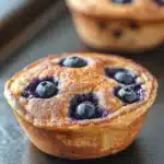 Blueberry muffin pancake bowl - the image shows a close-up of a freshly baked blueberry muffin on a black baking tray. the muffin has a golden brown crust and is filled with fresh blueberries. the blueberries are arranged in a circular pattern on top of the muffin, with some of them overlapping each other. the tray is lined with parchment paper and there is a white bowl in the background. the overall mood of the image is warm and inviting.