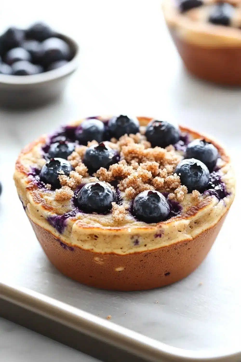 Blueberry muffin pancake bowl - the image shows a close-up of a blueberry tart on a baking tray. the tart has a golden brown crust and is topped with fresh blueberries. the blueberries are arranged in a crumbly manner on top of the tart, with some of them overlapping each other. in the background, there is a small bowl of more blueberries, suggesting that the tart is ready to be served. the tray is on a white countertop.