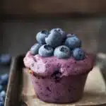 Blueberry muffin smoothie - the image is a close-up of a blueberry muffin on a wooden tray. the muffin is a light purple color and has a smooth texture. on top of the muffin, there are fresh blueberries arranged in a circular pattern. the blueberries are a deep blue color and appear to be ripe and juicy. the tray is lined with parchment paper and there are a few more blueberries scattered around it. the background is blurred, but it appears to be a rustic wooden table. the overall mood of the image is fresh and appetizing.