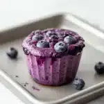 Blueberry muffin smoothie - the image shows a freshly baked blueberry muffin on a baking tray. the muffin is a deep purple color and is topped with fresh blueberries. the tray is lined with parchment paper and there are a few scattered blueberries around the muffin. the background is a white marble countertop.