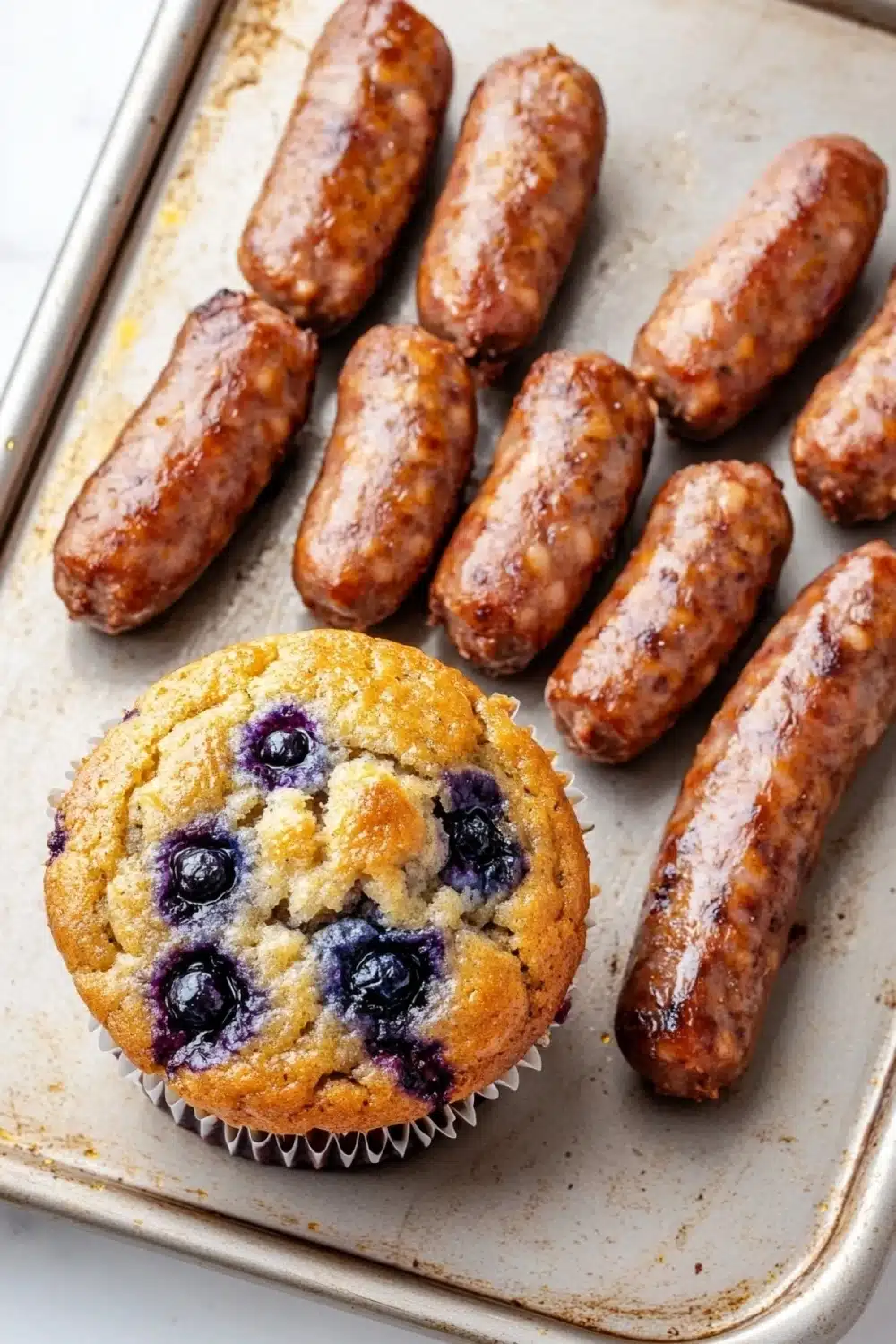 Blueberry muffin with sausage - the image shows a muffin on a baking tray. the muffin is golden brown and has a crumbly texture. it is topped with fresh blueberries. on the tray, there are several small sausages arranged in a single row. the sauses appear to be cooked and have a golden brown color. the tray is lined with parchment paper.