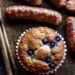Blueberry muffin with sausage - the image shows a muffin in a white paper liner on a baking tray. the muffin is golden brown and has a crumbly texture. it is filled with blueberries and appears to be freshly baked. there are several sausages scattered around the tray, some of which are whole and some are cut in half. the tray is lined with parchment paper and there is a black countertop in the background.