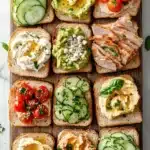 Bread toppings - the image is a flat lay of a wooden platter with nine slices of bread arranged in a grid-like pattern. each slice has a different type of avocado spread on it. the avocado spread is a bright green color and appears to be freshly made. on top of the avocado spread, there are slices of sliced cucumber, cherry tomatoes, and a dollop of sour cream. on the right side of the platter, there is a piece of grilled chicken with a sprinkle of herbs on top. the platter is garnished with fresh basil leaves. the background is a white marble countertop.