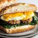 Breakfast sandwich ciabatta - the image shows a breakfast sandwich on a plate. the sandwich is made with a toasted bun and is topped with a fried egg on top. the egg is cooked sunny-side up and is oozing out of the sides. the sausage is cooked to a golden brown and appears to be seasoned with herbs and spices. the plate is gray with a speckled pattern and there is another sandwich visible in the background.