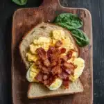 Breakfast sandwich on sourdough bread - the image shows a wooden cutting board with a sandwich on it. the sandwich is made with two slices of toasted bread, topped with scrambled eggs and bacon. the eggs are yellow and runny, and the bacon is crispy and golden brown. there are two sprigs of fresh spinach on the cutting board, adding a pop of green color to the dish. the cutting board is placed on a dark wooden table.