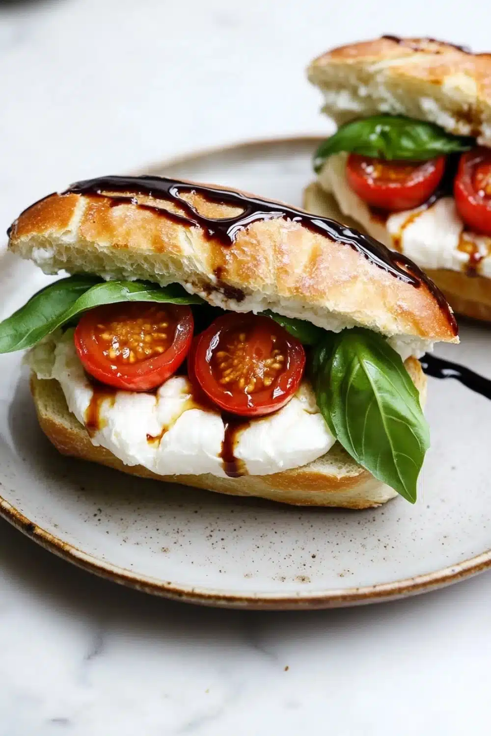 Burrata caprese sandwich with tomato confit - the image shows two sandwiches on a white plate. the sandwiches are made with a toasted bread and are topped with a generous amount of mozzarella cheese, sliced cherry tomatoes, and fresh basil leaves. the cheese is melted and bubbly, and the tomatoes are bright red. the basil leaves are a vibrant green color and are arranged in a circular pattern around the sandwich. the plate is sitting on a marble countertop. the background is blurred, but it appears to be a kitchen countertop with other plates and utensils visible.