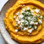 Butternut squash whipped feta dip - the image is a close-up of a bowl of mashed potatoes. the mashed potatoes are a bright orange color and appear to be smooth and creamy. on top of the mashed potatoes, there is a dollop of feta cheese and a sprig of dill. the bowl is made of ceramic with a speckled pattern around the edges. the background is a white marble countertop.