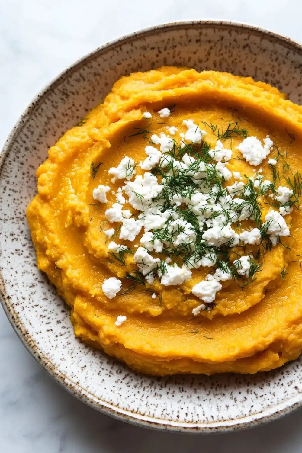 Butternut squash whipped feta dip - the image is a close-up of a bowl of mashed potatoes. the mashed potatoes are a bright orange color and appear to be smooth and creamy. on top of the mashed potatoes, there is a dollop of feta cheese and a sprig of dill. the bowl is made of ceramic with a speckled pattern around the edges. the background is a white marble countertop.