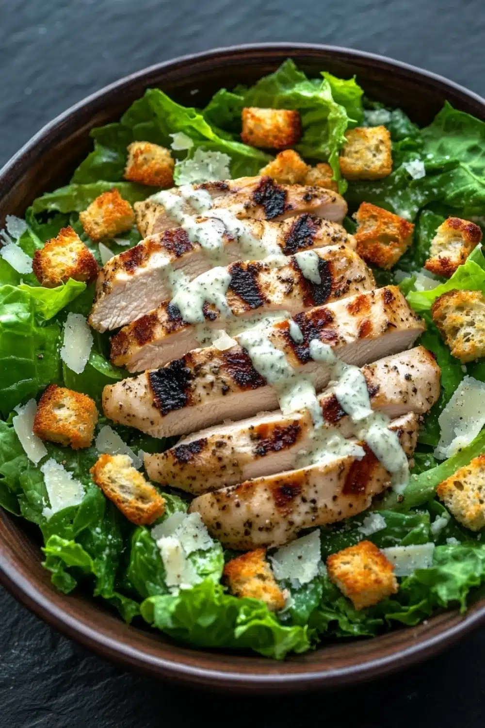 Caesar salad over chicken - the image is a close-up of a salad in a brown bowl. the salad is made up of green lettuce leaves and is topped with grilled chicken strips, croutons, and a drizzle of white sauce. the chicken strips are arranged in a circular pattern and appear to be seasoned with herbs and spices. the sauce is drizzled over the top of the chicken strips. the bowl is sitting on a dark grey countertop.