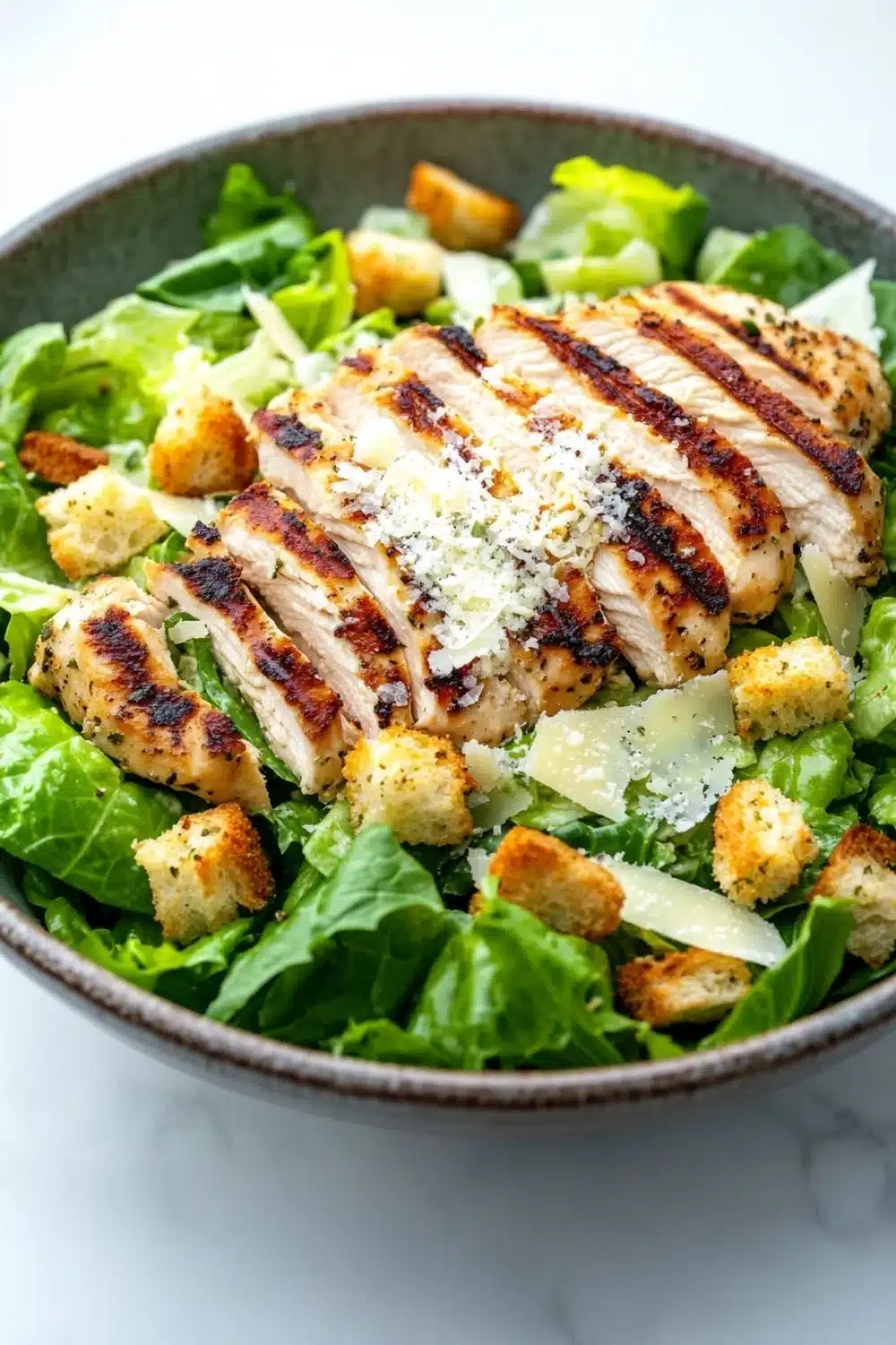 Caesar salad over chicken - the image is a close-up of a salad in a bowl. the salad is made up of green leafy vegetables, possibly romaine lettuce, and is topped with grilled chicken breasts. the chicken is cooked to a golden brown color and is drizzled with grated parmesan cheese. there are also croutons scattered throughout the salad, adding a crunchy texture. the bowl is sitting on a white marble countertop.