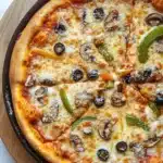 Cast iron pizza deep dish - the image is of a large pizza in a black cast iron skillet. the pizza is round and has a golden brown crust. it is topped with melted cheese, sliced mushrooms, green bell peppers, and black olives. the cheese is melted and bubbly, and the toppings are arranged in a circular pattern around the edges of the pizza. the skillet is sitting on a white marble countertop.
