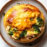 Cheesy chicken broccoli casserole buns in my oven - the image shows a round, golden-brown bread roll on a white marble countertop. the bread roll is topped with a layer of melted cheese and chunks of chicken and broccoli. the cheese is melted and bubbly, and the broccoli is bright green. the dish appears to be freshly baked and ready to eat.
