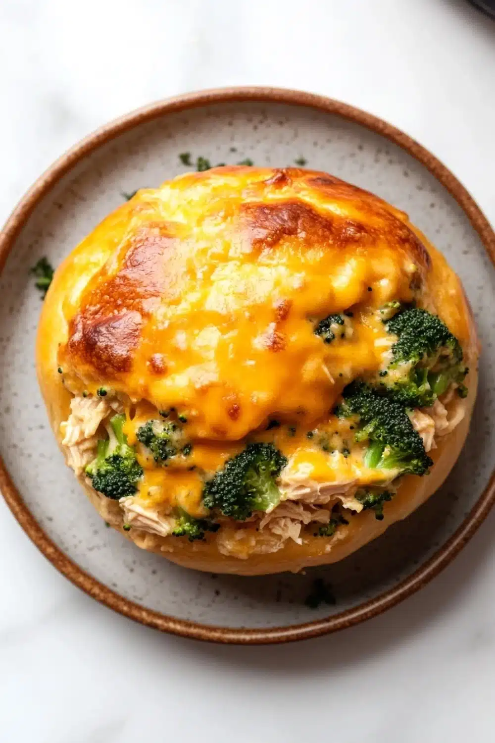 Cheesy chicken broccoli casserole buns in my oven - the image shows a round, golden-brown bread roll on a white marble countertop. the bread roll is topped with a layer of melted cheese and chunks of chicken and broccoli. the cheese is melted and bubbly, and the broccoli is bright green. the dish appears to be freshly baked and ready to eat.