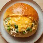 Cheesy chicken broccoli casserole buns in my oven - the image shows a round plate with a sandwich on it. the sandwich is made with a golden brown bun and is topped with melted cheese and broccoli. the cheese is yellow and appears to be melted and bubbly. the broccoli is cut into small pieces and is scattered throughout the sandwich. the plate is white with a light brown speckled pattern. the background is a wooden table.