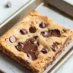 Chewy chocolate chip cookie bars - the image shows a square-shaped cookie bar on a baking tray. the cookie bar is golden brown in color and has a crumbly texture. it is topped with chocolate chips and drizzled with a thick layer of chocolate sauce. the tray is lined with parchment paper and there are scattered chocolate chips around the tray. in the background, there is a white marble countertop.