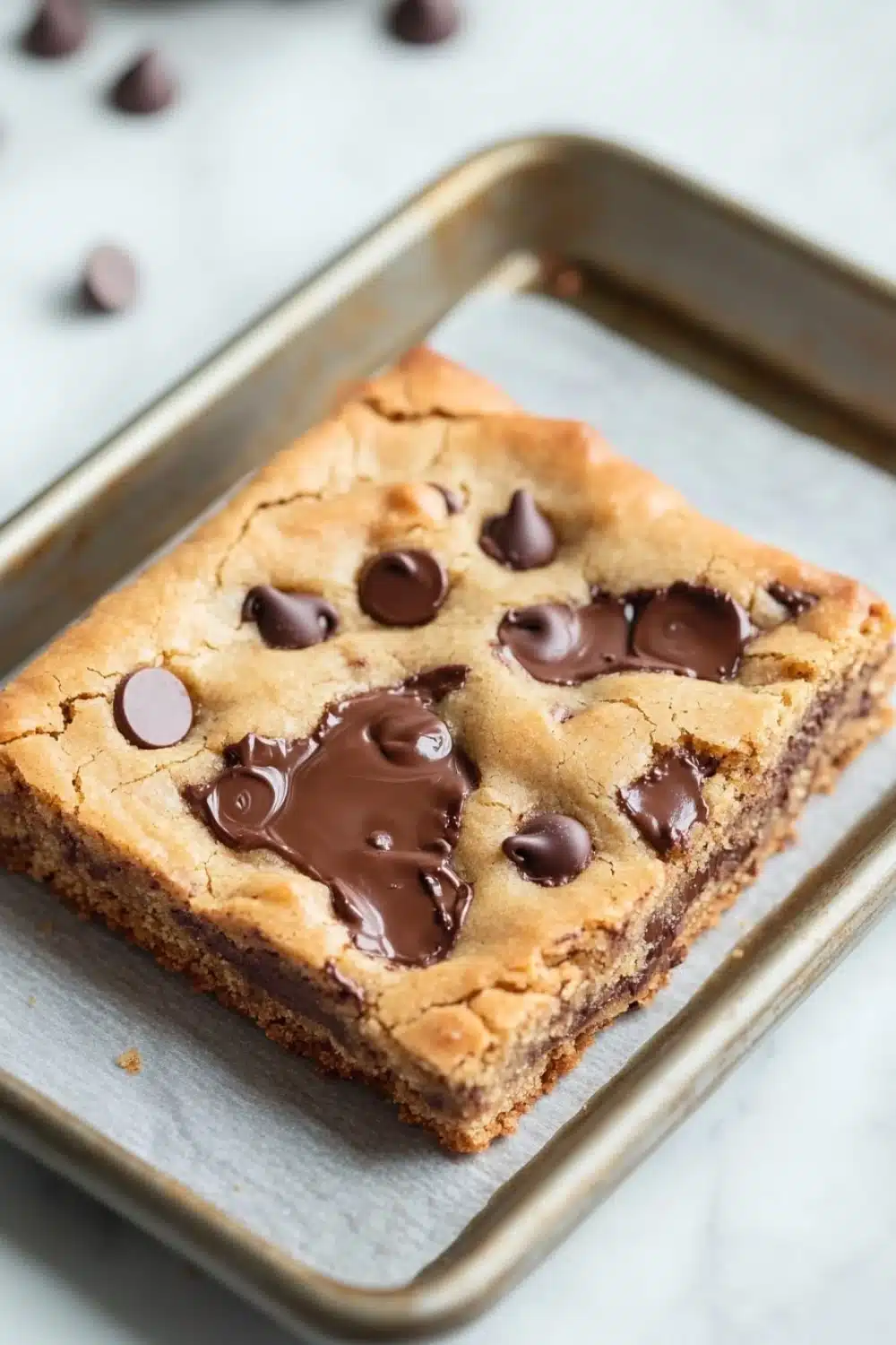 Chewy chocolate chip cookie bars - the image shows a square-shaped cookie bar on a baking tray. the cookie bar is golden brown in color and has a crumbly texture. it is topped with chocolate chips and drizzled with a thick layer of chocolate sauce. the tray is lined with parchment paper and there are scattered chocolate chips around the tray. in the background, there is a white marble countertop.