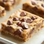 Chewy chocolate chip cookie bars - the image shows a close-up of a rectangular cookie bar on a baking tray. the cookie bar is golden brown in color and has a crumbly texture. it is topped with chocolate chips, giving it a shiny appearance. the tray is lined with parchment paper and there are other cookie bars visible in the background. the overall aesthetic of the image is rustic and appetizing.