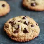 Chewy cookie - the image shows a group of freshly baked chocolate chip cookies on a dark grey surface. the cookies are round and golden brown in color, with chocolate chips scattered throughout. they are arranged in a scattered manner, with some overlapping each other. the background is blurred, but it appears to be a kitchen countertop. the overall mood of the image is warm and inviting.