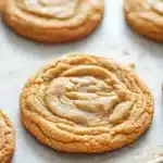 Chewy cookie base - the image shows a close-up of freshly baked peanut butter cookies on a baking sheet. the cookies are round and golden brown in color, with a swirl pattern on top. they are arranged in a single layer on the baking sheet, with some overlapping each other. the background is blurred, but it appears to be a wooden table or countertop. the overall mood of the image is warm and inviting.