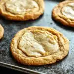 Chewy cookie base - the image shows a baking tray with six freshly baked cookies on it. the cookies are round and golden brown in color, with a crumbly texture on top. they are arranged in a row on the tray, with some overlapping each other. the tray is covered in a light dusting of powdered sugar, indicating that the cookies have been freshly baked. the background is dark, making the cookies stand out even more.