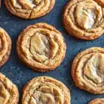 Chewy peanut butter cookie - the image shows a tray of freshly baked peanut butter cookies. the cookies are round and golden brown in color, with a crumbly texture on top. they are arranged in a circular pattern on a dark blue baking tray. some of the cookies have a swirl pattern on top, while others have a more rounded shape. the peanut butter filling is visible in the center of each cookie, creating a smooth and creamy texture. the tray appears to be lined with parchment paper.