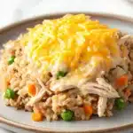 Chicken and brown rice casserole - the image shows a plate of fried rice with shredded chicken and peas. the rice appears to be cooked and has a golden brown color. the chicken is cut into small pieces and is covered in a layer of melted cheese. the peas are bright green and are scattered throughout the dish. the dish is served on a light blue plate with a white marble countertop in the background.