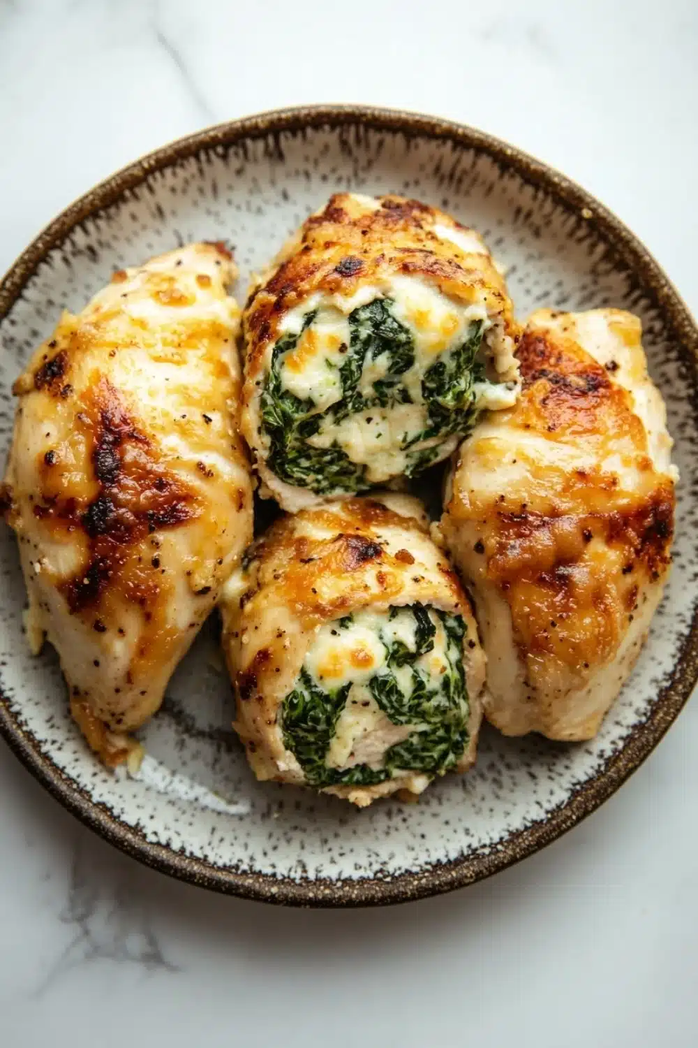 Chicken breast roll ups - the image shows a plate of four pieces of chicken breasts stuffed with spinach and cheese. the chicken breasts are golden brown and appear to be seasoned with herbs and spices. the cheese is melted and bubbly on top, and the spinach is a vibrant green color. the plate is round and has a speckled pattern around the edges. the background is a white marble countertop.