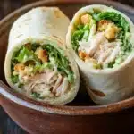 Chicken caesar salad pinwheels - the image shows two wraps in a brown ceramic bowl. the wraps are rolled up and placed on a wooden table. the wrap on the left is filled with shredded chicken, crumbled breadcrumbs, and a bed of green lettuce. the chicken appears to be cooked and has a golden brown color. the lettuce is a vibrant green color and looks fresh and healthy. the bowl is sitting on a dark wooden surface.