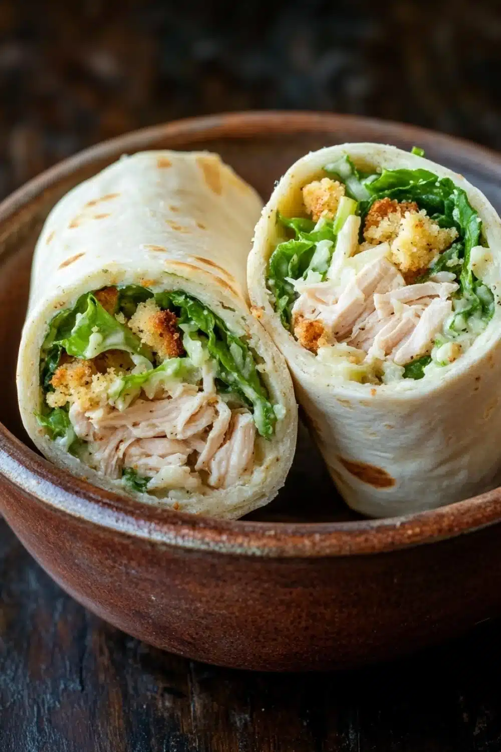 Chicken caesar salad pinwheels - the image shows two wraps in a brown ceramic bowl. the wraps are rolled up and placed on a wooden table. the wrap on the left is filled with shredded chicken, crumbled breadcrumbs, and a bed of green lettuce. the chicken appears to be cooked and has a golden brown color. the lettuce is a vibrant green color and looks fresh and healthy. the bowl is sitting on a dark wooden surface.