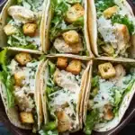 Chicken caesar salad tacos - the image shows a brown ceramic bowl filled with four tacos. the tacos are filled with shredded chicken, croutons, and a bed of green lettuce. the chicken appears to be seasoned with herbs and spices, and the crouton is sprinkled on top. the bowl is sitting on a dark grey countertop.