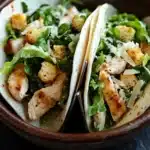Chicken caesar salad tacos - the image shows two tacos in a brown ceramic bowl. the tacos are filled with a salad of green arugula, croutons, and grilled chicken. the chicken is cooked to a golden brown color and appears to be seasoned with herbs and spices. the salad is garnished with grated parmesan cheese. the bowl is sitting on a dark grey countertop.