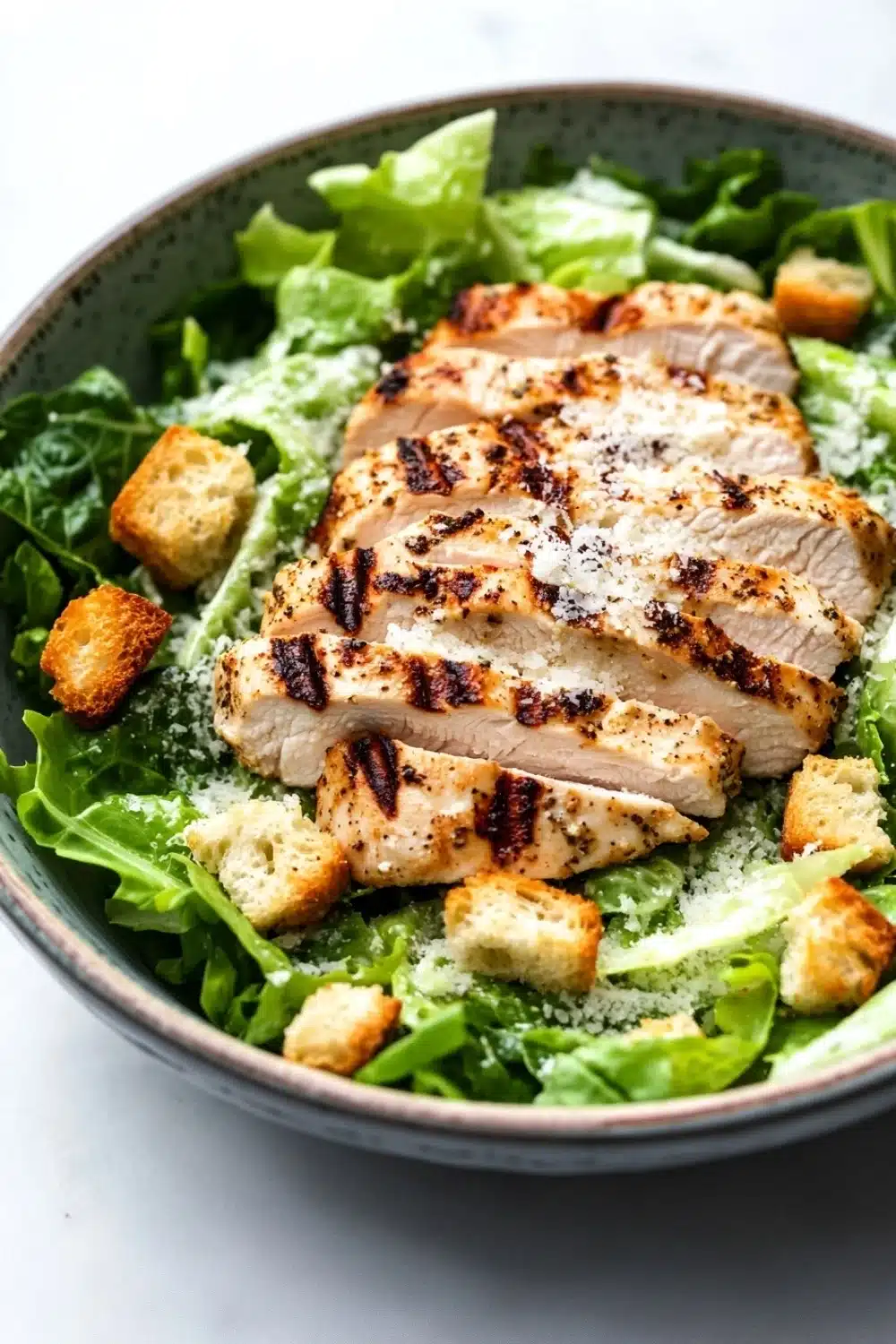 Chicken caesar salad - the image is a close-up of a salad in a blue bowl. the salad is made up of green leafy vegetables, possibly romaine lettuce, and is topped with grilled chicken strips. the chicken strips are arranged in a circular pattern and are drizzled with a light dressing. there are also croutons scattered throughout the salad, adding a crunchy texture. the bowl is sitting on a white countertop.