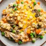 Chicken casserole creamy - the image is a close-up of a plate of food. the plate is round and has a light brown rim. the food appears to be a casserole dish with chunks of chicken, vegetables, and crumbled cheese. the chicken is cooked until it is golden brown and the vegetables include carrots, peas, and mushrooms. the cheese is melted and bubbly on top. the dish is garnished with a sprinkle of herbs and spices. the background is a white wooden table.