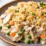 Chicken casserole creamy - the image is a close-up of a plate of food. the dish appears to be a creamy white sauce with chunks of chicken, peas, carrots, and other vegetables mixed in. the top of the dish is covered in a crumbly, golden-brown breadcrumbs, which are sprinkled with chopped parsley. the plate is brown and has a textured surface. the background is blurred, but it seems to be an indoor setting with a white tablecloth.