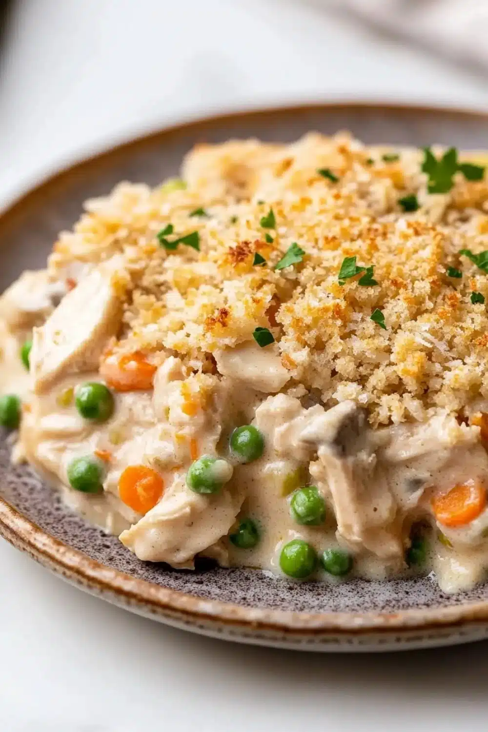 Chicken casserole creamy - the image is a close-up of a plate of food. the dish appears to be a creamy white sauce with chunks of chicken, peas, carrots, and other vegetables mixed in. the top of the dish is covered in a crumbly, golden-brown breadcrumbs, which are sprinkled with chopped parsley. the plate is brown and has a textured surface. the background is blurred, but it seems to be an indoor setting with a white tablecloth.