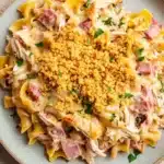 Chicken cordon blue freezer casserole - the image is a close-up of a plate of pasta dish. the pasta appears to be fusilli, which is a type of penne pasta, and it is topped with chunks of chicken, diced ham, and a creamy sauce. the dish is garnished with chopped parsley and a sprinkle of crumbled breadcrumbs. the plate is light blue in color and is sitting on a white countertop.