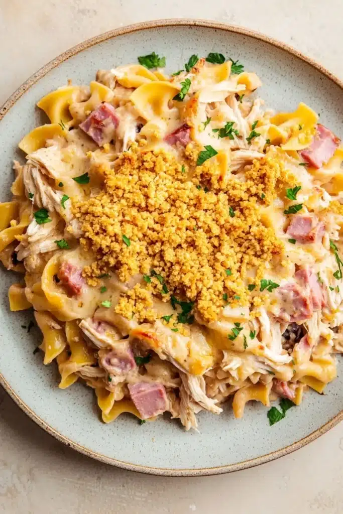 Chicken cordon blue freezer casserole - the image is a close-up of a plate of pasta dish. the pasta appears to be fusilli, which is a type of penne pasta, and it is topped with chunks of chicken, diced ham, and a creamy sauce. the dish is garnished with chopped parsley and a sprinkle of crumbled breadcrumbs. the plate is light blue in color and is sitting on a white countertop.