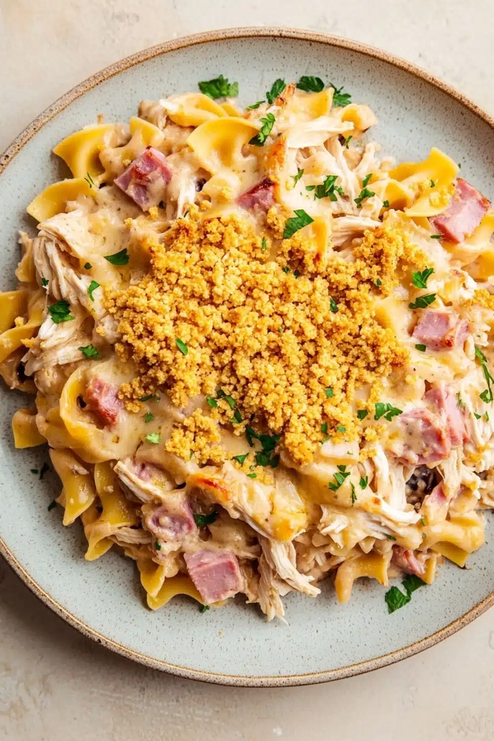 Chicken cordon blue freezer casserole - the image is a close-up of a plate of pasta dish. the pasta appears to be fusilli, which is a type of penne pasta, and it is topped with chunks of chicken, diced ham, and a creamy sauce. the dish is garnished with chopped parsley and a sprinkle of crumbled breadcrumbs. the plate is light blue in color and is sitting on a white countertop.