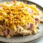 Chicken cordon blue freezer casserole - the image shows a plate of food, specifically a casserole dish. the dish appears to be made with shredded chicken, ham, and cheese, and is topped with a golden brown crumbly topping. the plate is gray with a speckled pattern and is sitting on a white marble countertop. there are a few sprigs of parsley scattered around the plate.