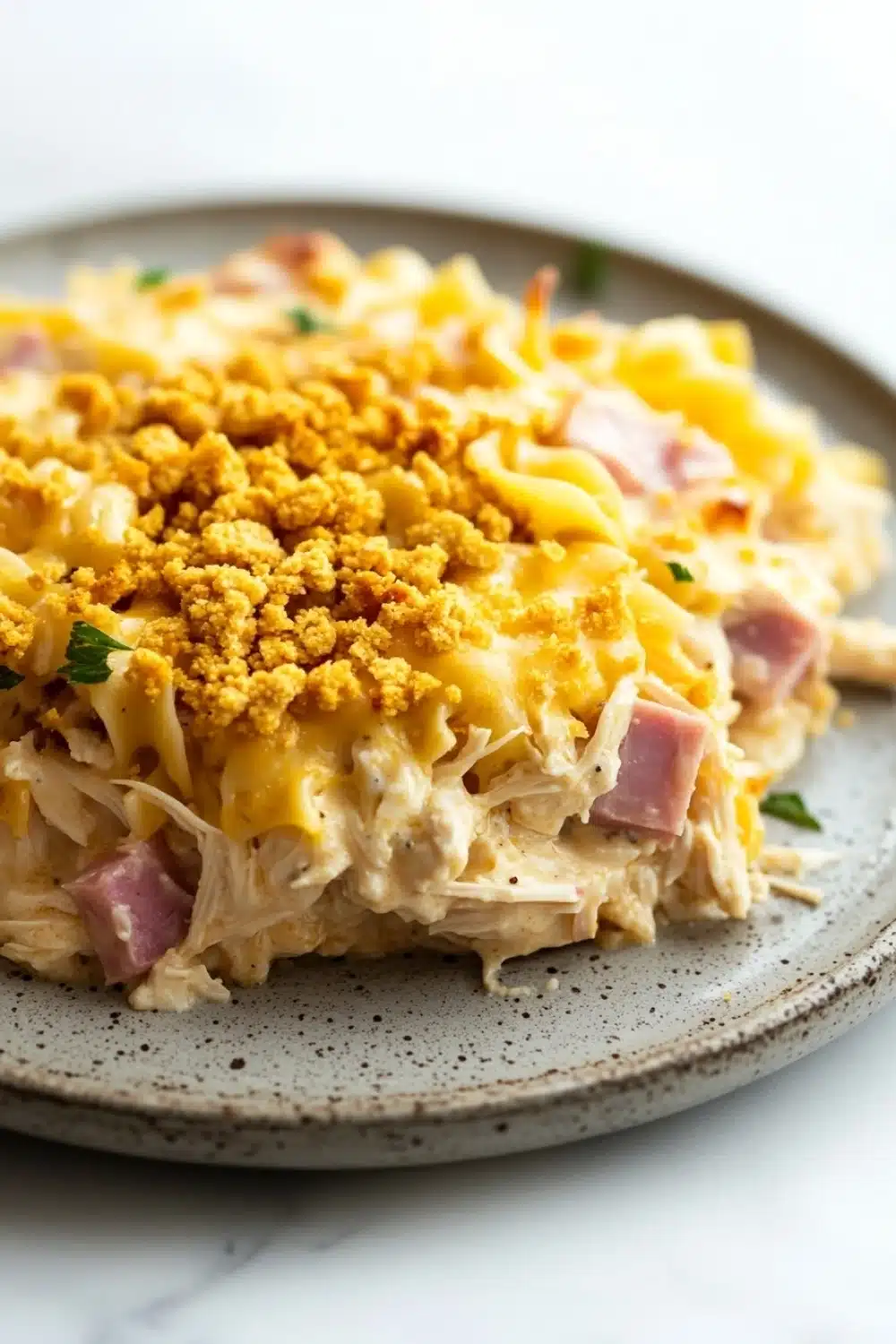 Chicken cordon blue freezer casserole - the image shows a plate of food, specifically a casserole dish. the dish appears to be made with shredded chicken, ham, and cheese, and is topped with a golden brown crumbly topping. the plate is gray with a speckled pattern and is sitting on a white marble countertop. there are a few sprigs of parsley scattered around the plate.