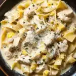 Chicken noodle skillet with sauce - the image is a close-up of a bowl of pasta dish. the pasta appears to be fettuccine alfredo, with chunks of chicken and yellow bell peppers mixed in. the dish is garnished with grated parmesan cheese and chopped herbs. the bowl is made of dark brown ceramic and is sitting on a wooden table. the background is blurred, so the focus is on the dish.