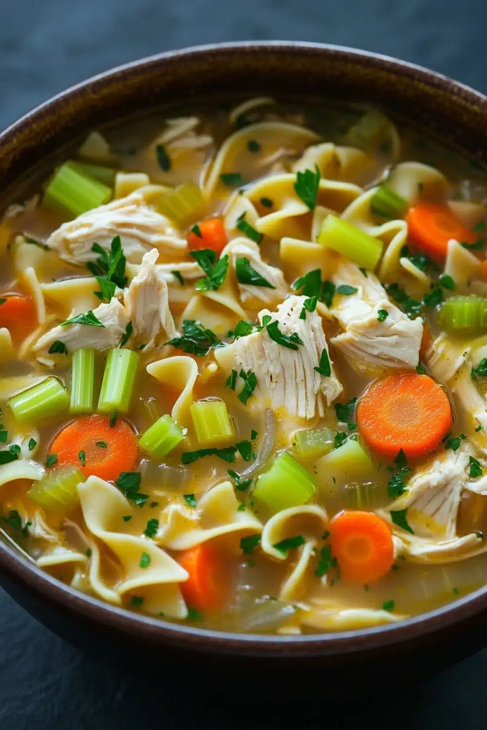 Chicken noodle soup in instant pot - the image is a close-up of a bowl of soup. the soup appears to be a type of noodle soup with chunks of chicken, carrots, celery, and other vegetables mixed in. the broth is a light yellow color and is thick and creamy. the bowl is made of dark brown ceramic and is sitting on a dark blue surface. there are also some sprigs of parsley scattered throughout the soup, adding a pop of green color to the dish.