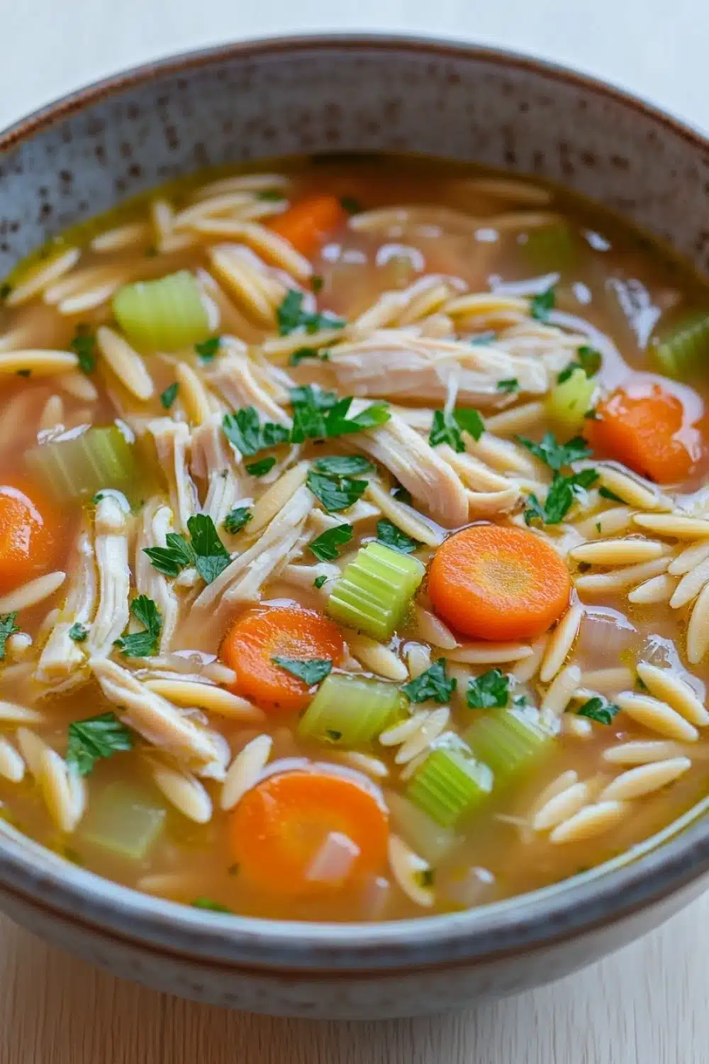 Chicken noodle soup with orzo - the image is a close-up of a bowl of soup. the soup appears to be a type of noodle soup with chunks of chicken, carrots, celery, and other vegetables mixed in. the bowl is made of ceramic and has a brown rim. the broth is a light brown color and looks thick and creamy. there are also some sprigs of parsley scattered throughout the soup, adding a pop of green color to the dish. the background is a white tablecloth.