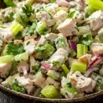 Chicken salad finely chopped - the image is a close-up of a bowl of salad. the salad appears to be made with diced chicken, celery, red onions, and cilantro. the chicken is cut into small cubes and is mixed with the celery and onions. the celery is bright green and the onions are red. the cilantro is scattered throughout the salad, adding a pop of green color to the dish. the bowl is made of dark wood and is sitting on a wooden table.