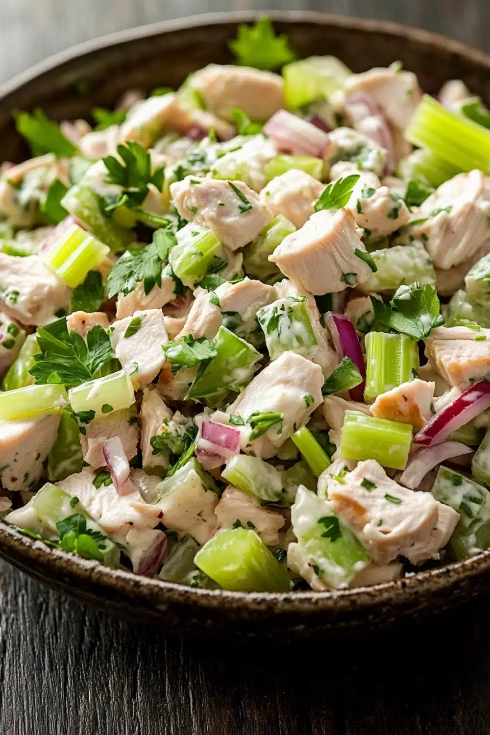 Chicken salad finely chopped - the image is a close-up of a bowl of salad. the salad appears to be made with diced chicken, celery, red onions, and cilantro. the chicken is cut into small cubes and is mixed with the celery and onions. the celery is bright green and the onions are red. the cilantro is scattered throughout the salad, adding a pop of green color to the dish. the bowl is made of dark wood and is sitting on a wooden table.