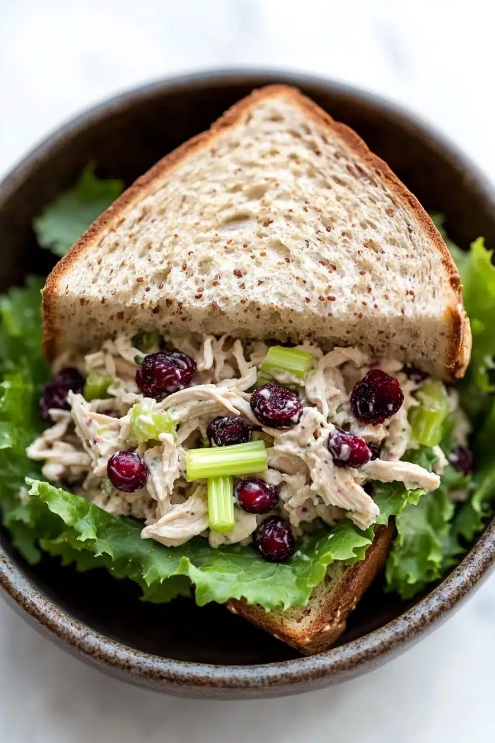 Chicken salad sandwich with cranberries - the image shows a bowl of salad with a slice of bread on top. the salad appears to be made with chicken, celery, and cranberries. the bowl is made of dark brown ceramic and is sitting on a white marble countertop. the bread is toasted and has a golden brown crust. the cranberries are bright red and appear to be ripe and juicy. the celery is a light green color and is scattered throughout the salad. the overall appearance of the salad is fresh and appetizing.