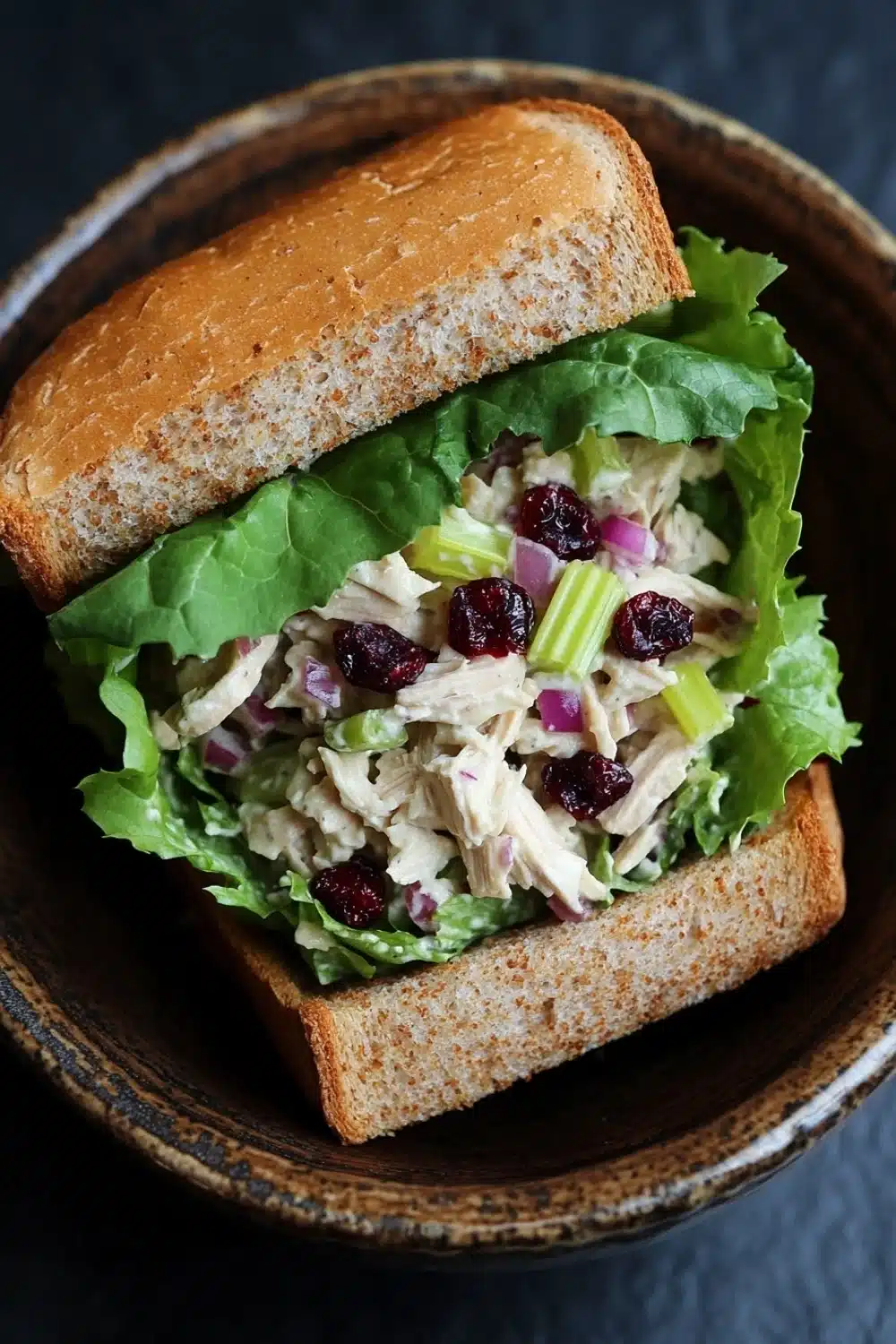 Chicken salad sandwich with cranberries - the image shows a sandwich in a brown ceramic bowl. the sandwich is made with a toasted bun and is filled with a mixture of lettuce, chicken, cranberries, celery, and other vegetables. the lettuce is a vibrant green color and the chicken is a light brown color. the cranberries are a deep red color and are scattered throughout the salad. the celery is a bright green and appears to be celery. the bowl is sitting on a dark grey countertop.