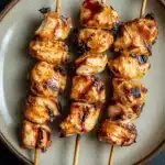 Chicken satay skewers appetizers - the image shows a plate of grilled chicken skewers. the skewers are arranged in a circular pattern on the plate, with each skewer slightly overlapping the one below it. the chicken appears to be seasoned with herbs and spices, giving it a golden brown color. the plate is light brown in color and has a textured surface. the background is dark, making the skewers stand out.